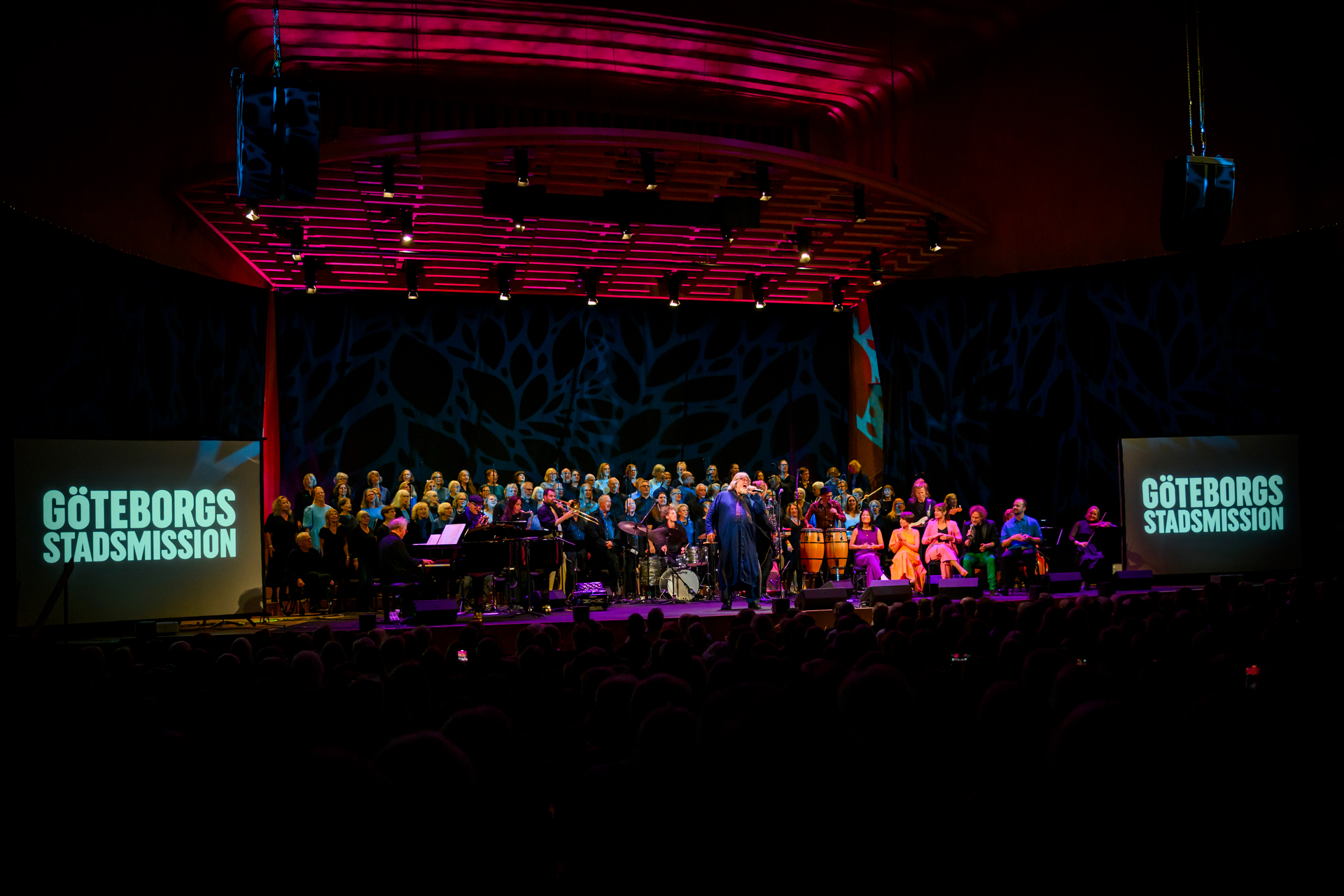 En stor kör och artister på Göteborgs konserthus stora scen samt tv-skärmar på båda sidor av scenen där det står Göteborgs Stadsmission.