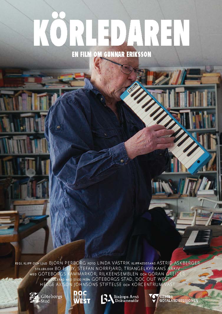 En man står framför en bokhyllevägg och blåser i en blå harmonica.