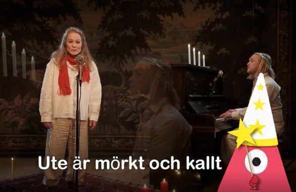 En kvinna som står framför en mikrofon och sjunger, en man som spelar piano bredvid i en gammaldags miljö med ljusstakar med levande ljus.